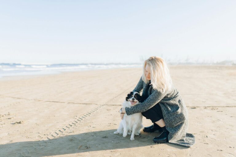 A woman in a long coat hugs her dog on a sunny beach, enjoying a peaceful day.