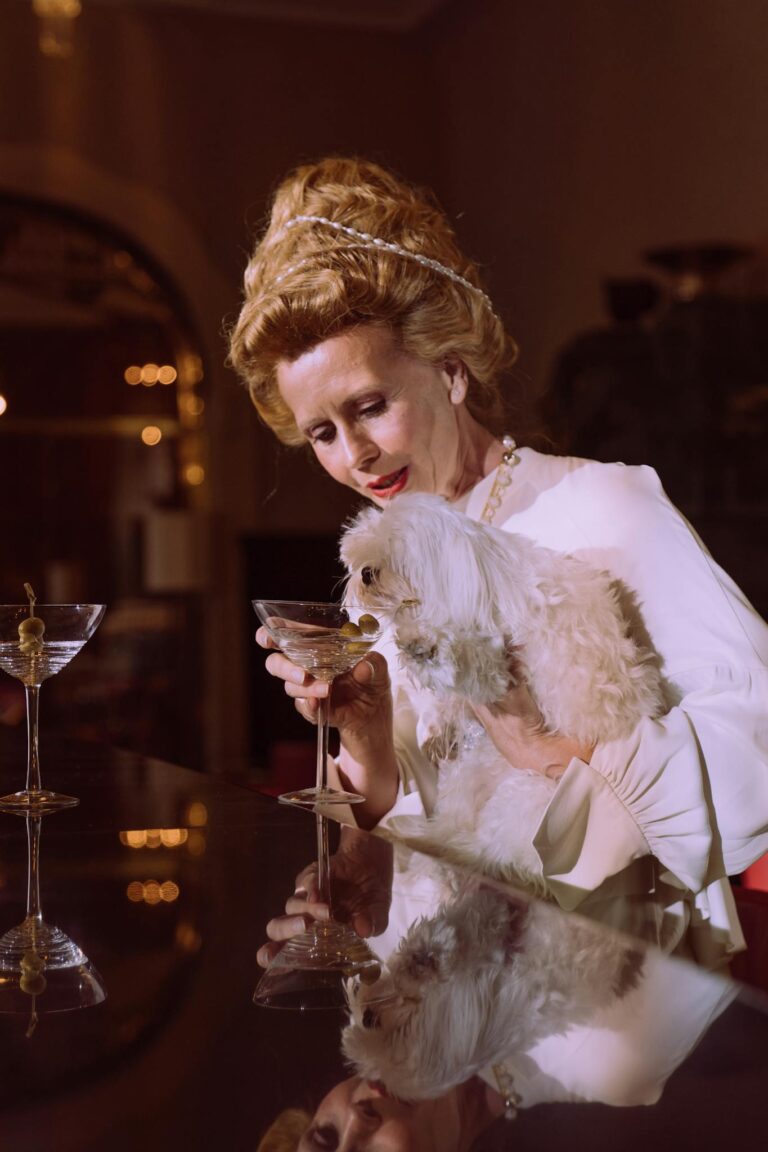 Sophisticated woman in elegant attire enjoys a drink with her Maltese dog at a stylish indoor bar.